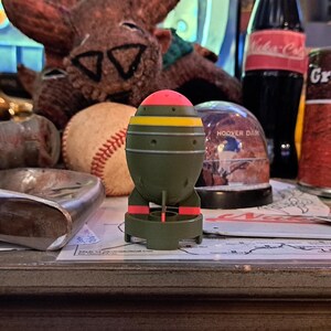Op de afbeelding: Een groene speelgoedbom met een roze bovenkant en gele strepen en rode accenten. Het speelgoed staat op een kaart, met een honkbal, een sneeuwbol en een conservenblik op de achtergrond.