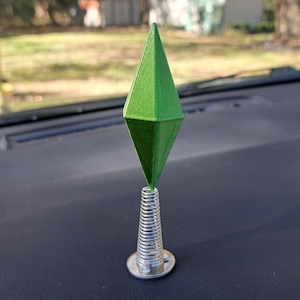May include: A green, diamond-shaped object, reminiscent of a video game icon, is mounted on a silver spring and base. The object is set against a dark dashboard, with a blurred outdoor background.