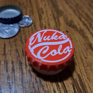 Puede incluir: Una chapa de botella Nuka Cola roja y blanca. La chapa tiene las palabras "Nuka Cola" en una fuente blanca estilizada. Otras chapas de botella son visibles en el fondo.