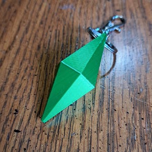 Pode incluir: Um chaveiro em forma de diamante verde brilhante com fecho e argola prateados. O berloque tem um design geométrico com múltiplas facetas. O chaveiro está sobre uma superfície de madeira.