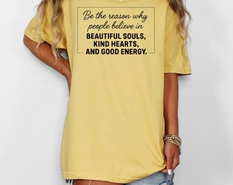 May include: Mustard yellow t-shirt with the text "Be the reason why people believe in BEAUTIFUL SOULS, KIND HEARTS, AND GOOD ENERGY" in a black rectangular frame. The shirt is a relaxed fit.