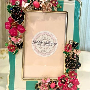 May include: A rectangular picture frame with a teal and white striped border, decorated with metal floral and butterfly embellishments in shades of pink, black, and green. A circular sticker in the center displays the text Gilded Whimsy VINTAGE DESIGN.