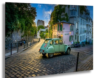 Op de afbeelding: Een lichtgroene oldtimer geparkeerd op een geplaveide straat voor een roze gebouw met groene luiken. De scène speelt zich af in een Europese stad met weelderig groen en een blauwe lucht.