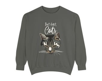 May include: A dark gray sweatshirt featuring a cat face graphic with the text "But first, Cats" in white. The cat graphic is a detailed illustration with shades of gray, black, and white. The sweatshirt has long sleeves and a crew neck.