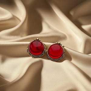 May include: A pair of vintage-style earrings with large, round, red cabochon stones set in ornate, gold-toned metal frames. The earrings are displayed on a backdrop of flowing, golden-colored fabric, creating a luxurious aesthetic.