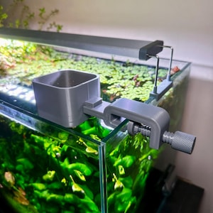 Può includere: Un supporto grigio per piante da acquario e un morsetto per l'apparecchio di illuminazione. Il morsetto è attaccato al vetro di un acquario pieno di piante verdi. Il portapiante è un contenitore quadrato. L'apparecchio di illuminazione è di forma lunga e rettangolare.