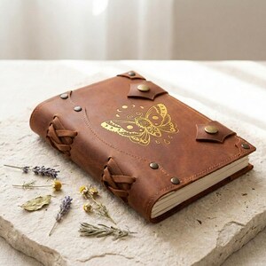 May include: A brown leather journal featuring a gold embossed butterfly design on the front cover. The journal is secured with metal studs and decorative leather lacing along the spine and edges, with visible cream-colored paper pages inside.