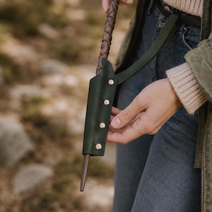 May include: A dark green leather holster featuring four metallic rivets and a matching strap, designed to hold a brown wooden wand with a textured, wrapped handle. The holster is constructed from smooth, matte leather and secures the wand vertically.