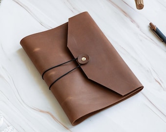May include: A brown leather journal cover with a pointed flap, secured by a single brass-colored metal snap button and a thin black elastic cord that wraps around the exterior. The leather has a smooth, matte finish with visible natural grain.