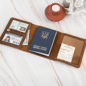 May include: Open brown leather wallet with a blue passport, currency, and identification cards. The wallet is open, displaying its contents against a marble surface. A small, round, terracotta candle holder and a white coral decoration are in the background.