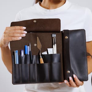Puede incluir: Estuche enrollable de artista de cuero marrón oscuro, abierto para mostrar los materiales de arte. El estuche tiene múltiples bolsillos que contienen bolígrafos, pinceles y una espátula. El estuche se mantiene abierto, revelando el contenido.