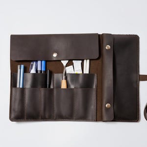 May include: A dark brown leather roll-up organizer featuring multiple vertical pockets containing various art supplies, including blue pens, a metal palette knife, paint tubes, and white pencils, secured with a long leather strap and metal snaps.