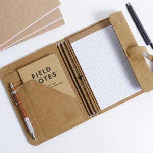 May include: A tan leather notebook cover featuring a pocket containing a Field Notes memo book, a grid-lined paper insert held by elastic cords, and a silver and wood-toned mechanical pencil. A black pen rests nearby on the surface.