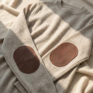 May include: A beige long-sleeve sweater featuring a crew neckline and ribbed cuffs. The garment is crafted from a textured knit fabric and includes smooth, oval-shaped brown patches stitched onto the exterior of both elbow areas.