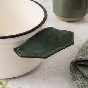 Może przedstawiać: Kremowa miska ceramiczna z ciemnym brzegiem i ciemnozielonym skórzanym uchwytem z wytłoczonymi literami AM. Misce towarzyszy pasujący zielony kubek oraz teksturowana zielona ściereczka, tworzące spójny i estetyczny zestaw przedmiotów.