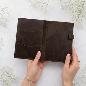 May include: A dark brown leather passport holder featuring two stacked chevron-shaped card slots on the left side, a larger vertical pocket on the right side, and a small leather loop for a pen attached to the right edge of the interior.