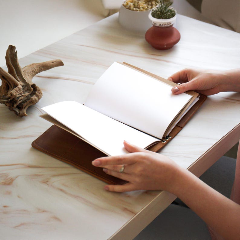 May include: A brown leather journal with blank white pages is open on a marble-patterned surface. The journal is held open by a person. A piece of driftwood and small potted plants are in the background.