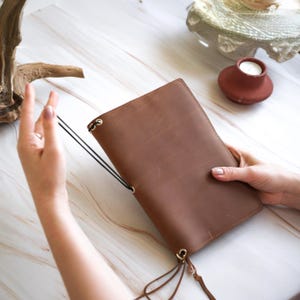 May include: A brown leather journal with a black elastic closure and gold-colored metal accents. The journal is held open, showcasing its interior pages. The leather has a textured appearance. The journal is approximately 8 inches tall and 5 inches wide.