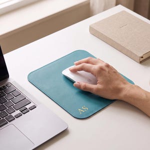 Op de afbeelding: Een vierkante teal leren muismat met afgeronde hoeken en gestikte randen. In de rechterbenedenhoek staan de in goud gedrukte initialen AS. Het oppervlak is glad en plat, ontworpen om een computermuis te ondersteunen tijdens dagelijks gebruik.