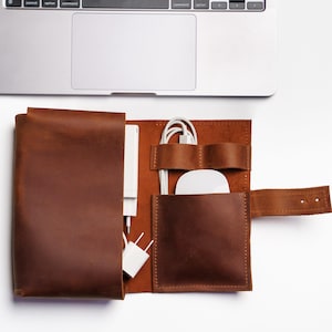 Puede incluir: Organizador tecnológico de cuero marrón abierto. Contiene un cargador blanco, un ratón blanco y un cable de carga blanco. Diseñado para guardar accesorios tecnológicos.