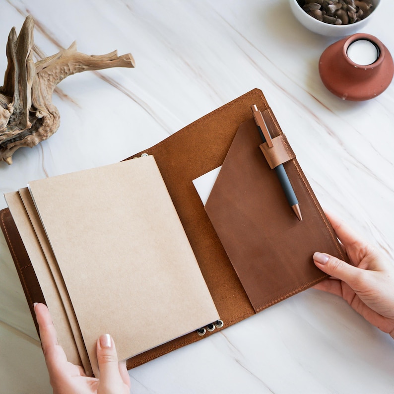 May include: Open brown leather journal with a pen in a holder. The journal has several blank pages and a pocket. The journal is open, revealing the inside pages. The pen is black with a copper-colored tip.