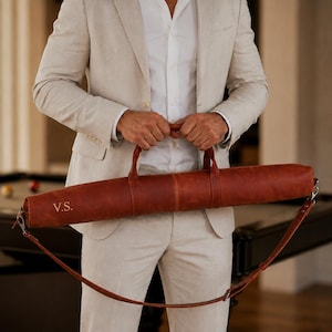 May include: A cylindrical brown leather pool cue case featuring two top handles and an adjustable shoulder strap. The exterior is embossed with the initials V.S. on the side. The case is constructed from smooth, matte-finish leather material.
