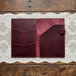 Può includere: Una copertina per taccuino in pelle color bordeaux scuro con finitura liscia sul lato sinistro, dotata di due fessure per carte a forma di chevron e una tasca interna testurizzata sul lato destro, completa di un piccolo passante portapenna.