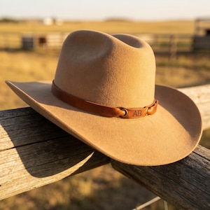 Könnte beinhalten: Ein beigefarbener Cowboyhut mit einem braunen Lederband und den Initialen "AB" auf dem Band. Der Hut liegt auf einem Holzzaun, mit einer verschwommenen ländlichen Landschaft im Hintergrund.