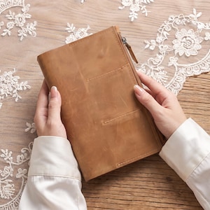 Puede incluir: Una funda de cuero color canela para cuaderno con un acabado mate suave y textura de grano natural visible. La funda incluye un cierre de cremallera lateral con una pequeña lengüeta de cuero y dos bolsillos cosidos en el frente.