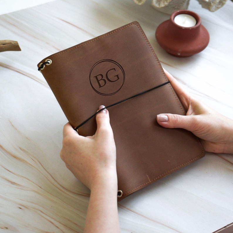 May include: A brown leather journal with the initials "BG" inside a circle. The journal is held closed with a black elastic band. The journal is a warm brown color.