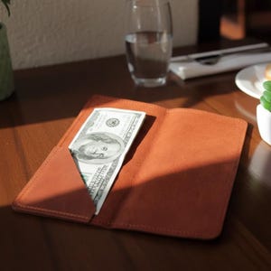 May include: A brown leather wallet with a hundred-dollar bill partially inserted. The wallet is open, revealing the bill's face. The scene is set on a wooden table, with a glass of water and a plate in the background.