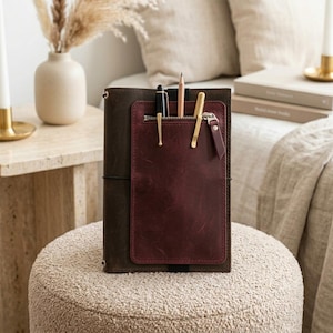 Könnte beinhalten: Ein dunkelbrauner Notizbuchumschlag aus Leder mit einer vertikalen bordeauxroten Ledertasche mit Reißverschlussfach. Die Tasche enthält einen schwarzen Füllfederhalter, einen Holzbleistift und einen goldfarbenen Metallstift. Ein schwarzes Gummiband sichert den Umschlag.