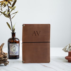 May include: A brown leather journal with the initials "AV" embossed on the cover. The journal is secured with a black elastic band and features a minimalist design. The background includes a decorative vase and dried foliage.