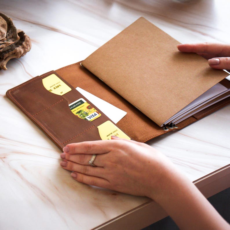 May include: A brown leather wallet with multiple card slots and a notebook. The wallet is open, revealing credit cards and a notepad. The notebook has a brown cover and white pages. The wallet is on a light-colored surface.