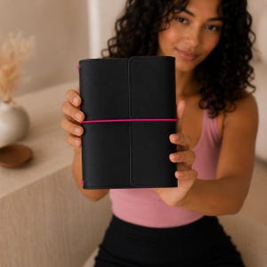 Könnte beinhalten: Ein schwarzes Lederjournal mit einem leuchtend pinkfarbenen Gummiband. Das Journal wird vor einer Person hochgehalten. Das Journal ist geschlossen und hat ein schlichtes, elegantes Design. Der Hintergrund ist neutral.