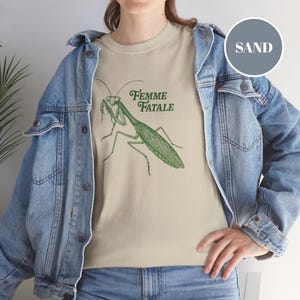 Puede incluir: Camiseta color arena con una ilustraci&oacute;n verde de una mantis religiosa y las palabras "Femme Fatale". El dise&ntilde;o est&aacute; impreso en una camiseta suave y de tono neutro.