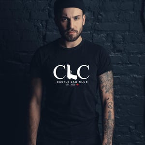Peut inclure: T-shirt noir avec le texte blanc "CLC", une silhouette de pistolet et "CASTLE LAW CLUB EST. 2025". Une petite flèche rouge vers le bas. Le t-shirt est porté par une personne.