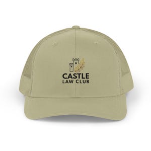 Op de afbeelding: Een beige truckerpet met een mesh achterkant en een gebogen klep. De voorkant toont de tekst "CASTLE LAW CLUB" onder een afbeelding van een kasteel en tarwe. De pet heeft een neutrale kleur.