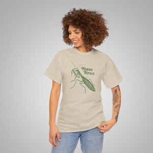 Puede incluir: Una camiseta beige con una ilustraci&oacute;n verde de una mantis religiosa y las palabras "Femme Fatale" en verde. La camiseta est&aacute; hecha de un material suave. La persona lleva vaqueros azules.