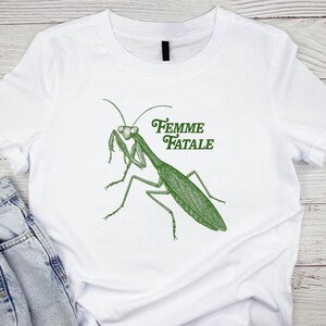 Puede incluir: Camiseta blanca con una ilustraci&oacute;n verde de una mantis religiosa y el texto "Femme Fatale". El dise&ntilde;o es un dibujo lineal detallado del insecto. La camiseta est&aacute; hecha de un material suave.
