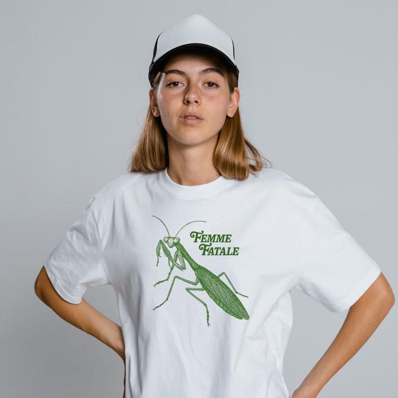 Puede incluir: Camiseta blanca con una ilustraci&oacute;n verde de una mantis religiosa y el texto "Femme Fatale". La persona lleva una gorra de camionero blanca y negra.