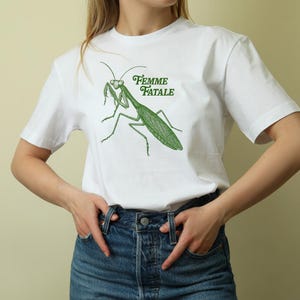 Puede incluir: Camiseta blanca con una ilustraci&oacute;n verde de una mantis religiosa y el texto "Femme Fatale". El dise&ntilde;o est&aacute; centrado en la parte delantera de la camiseta. La modelo lleva vaqueros azules.