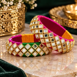 May include: Two circular bangles wrapped in vibrant pink, yellow, and green silk thread. Each bangle features a decorative band of square-shaped mirrors set in gold-toned metal frames arranged in a repeating diamond pattern around the exterior.
