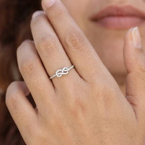 May include: A thin silver-toned ring featuring a central infinity knot design encrusted with small, clear, sparkling stones. The band is slender and metallic, showcasing a continuous, delicate loop pattern that sits horizontally across the finger.