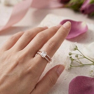 May include: A rose gold ring set featuring a central emerald-cut clear stone on a band accented with small pavé diamonds, paired with a matching thin pavé diamond band. The metal has a warm pinkish hue and the stones are clear and faceted.