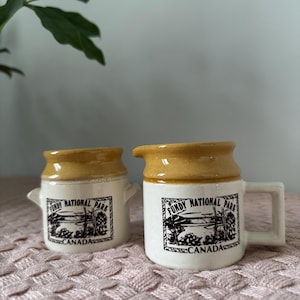 May include: Two small, vintage ceramic cream jugs. The jugs are white with a tan rim and handle. Each jug has a black and white graphic of Fundy National Park, Canada, with the text "Fundy National Park Canada".