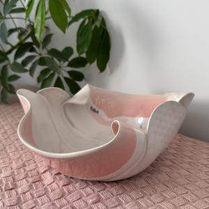 May include: A decorative ceramic bowl with a ruffled edge. The bowl has a white interior with a pink stripe and a pink exterior. The bowl is on a pink textured surface.