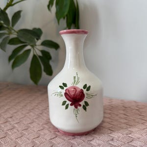 Puede incluir: Un jarrón de cerámica blanco con un diseño de rosa roja pintada a mano. El jarrón tiene un borde y una base rosas, y la rosa está rodeada de hojas verdes. Es un objeto decorativo.