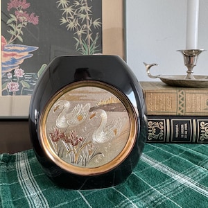 May include: A black vase with a gold rim and a decorative panel featuring two swans and floral accents. The vase is oval-shaped and sits on a green plaid cloth. A framed artwork and a candle holder are in the background.