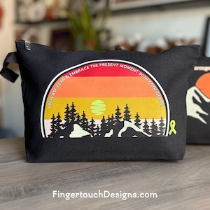May include: Black canvas zippered pouch with a colorful sunset design. The design includes a mountain range, trees, and the text "I RELEASE FEAR & EMBRACE THE PRESENT MOMENT WITH CONFIDENCE." A yellow ribbon is on the right side.